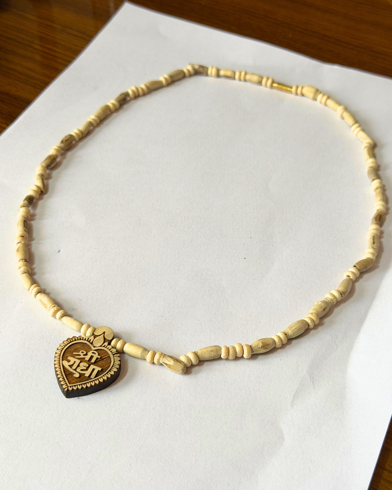 Cream-colored beaded necklace with a round carved wooden pendant placed on white paper. The pendant features a decorative carved design and text.