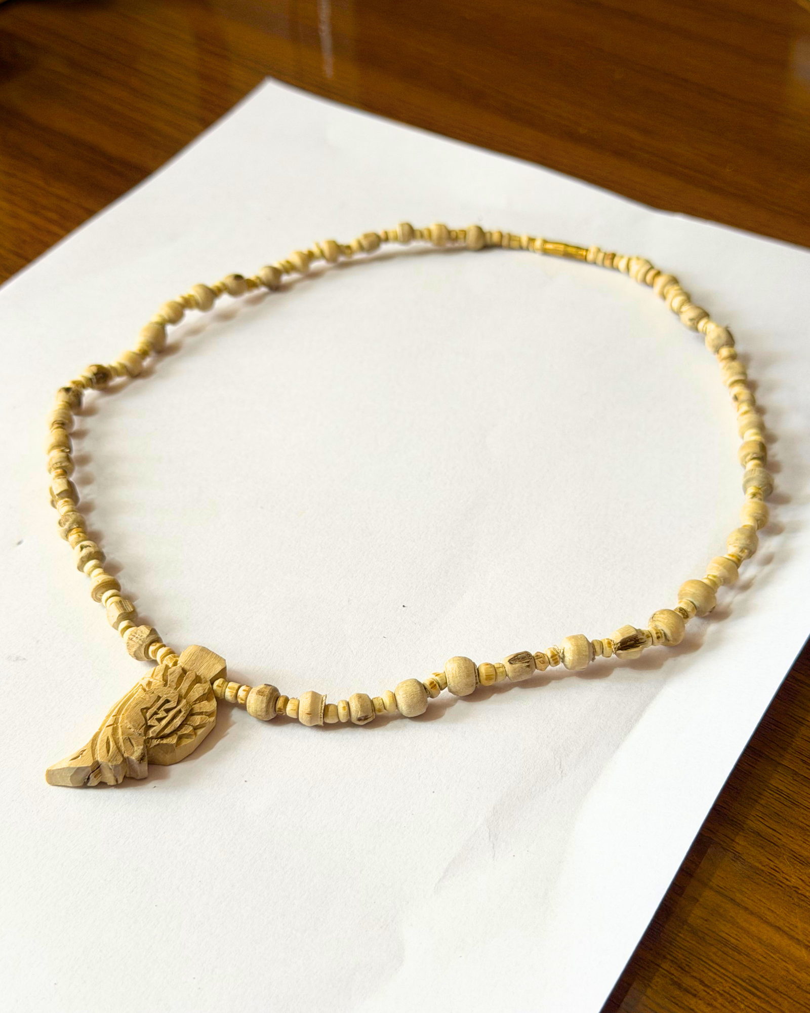 Cream-colored beaded necklace with a large carved pendant laid on white paper on a wooden table edge.