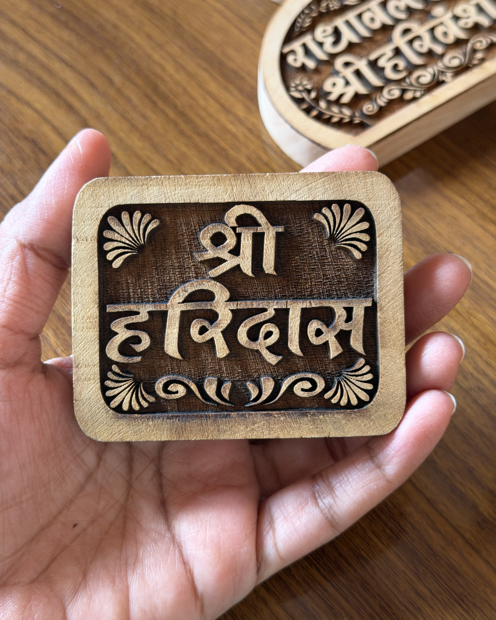 Close-up of a hand holding a carved wooden stamp with ornate Devanagari lettering and decorative flourishes on a wooden surface.